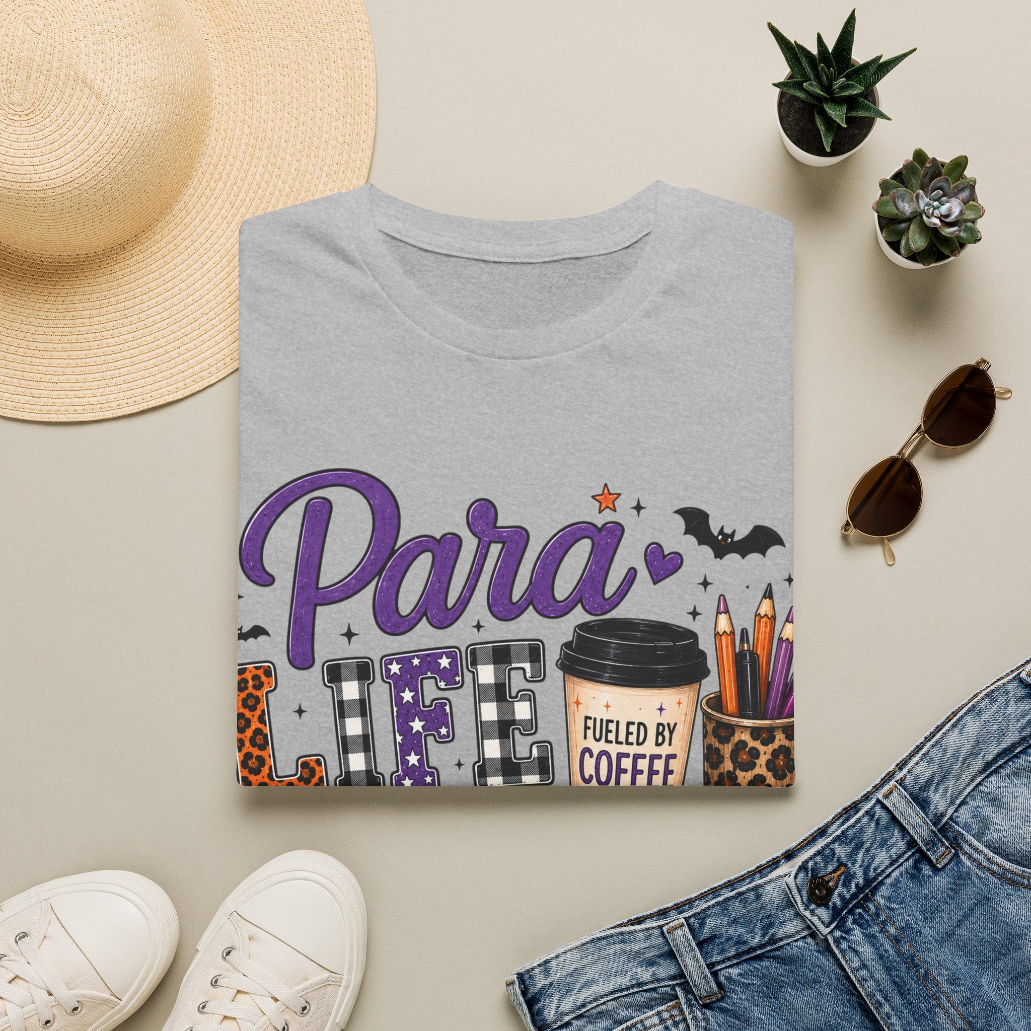 A folded light gray graphic T-shirt is styled in a flat lay on a beige surface with a straw hat, small potted succulents, sunglasses, white sneakers, and blue jeans around it. The shirt graphic reads 'Para LIFE' with a coffee cup and Halloween-themed details.