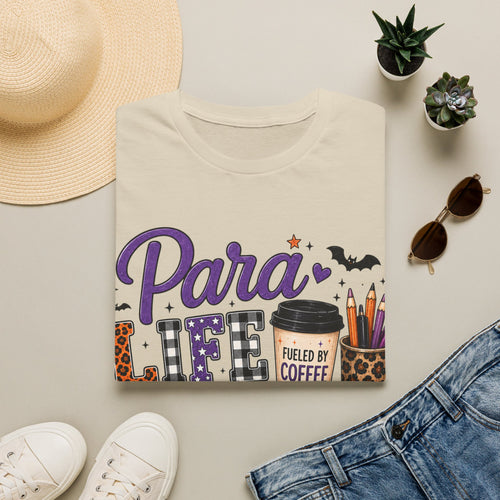 A folded cream-colored graphic T-shirt is styled in a flat lay with a straw hat, white sneakers, blue jeans, small potted succulents, and sunglasses on a light beige background. The shirt features a graphic that reads 'Para LIFE FUELED BY COFFEE' with a coffee cup, pencils, bats, and leopard print details.